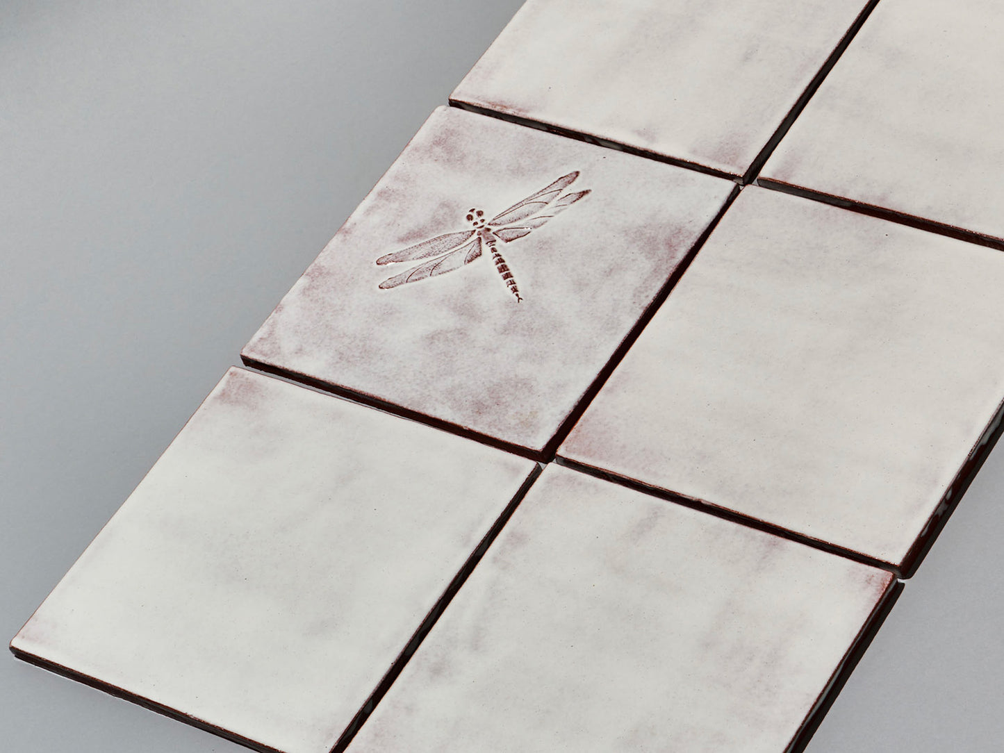 Quadratische, weiße Majolika-Fliese mit leichtem Verlauf und erhabenem Relief einer filigranen Libelle im Zentrum.
Square white-coloured majolica tile with subtle shading and a raised relief of a delicate damselfly in the centre.