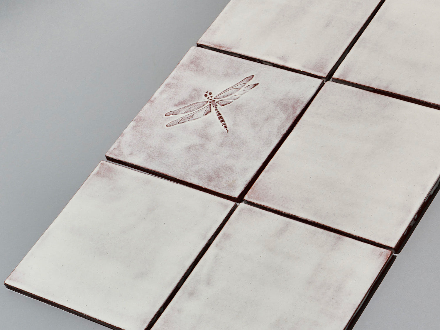Quadratische, weiße Majolika-Fliese mit leichtem Verlauf und erhabenem Relief einer filigranen Libelle im Zentrum.

Square white-coloured majolica tile with subtle shading and a raised relief of a delicate damselfly in the centre.