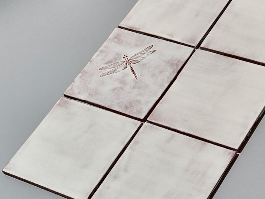 Quadratische, weiße Majolika-Fliese mit leichtem Verlauf und erhabenem Relief einer filigranen Libelle im Zentrum.

Square white-coloured majolica tile with subtle shading and a raised relief of a delicate damselfly in the centre.