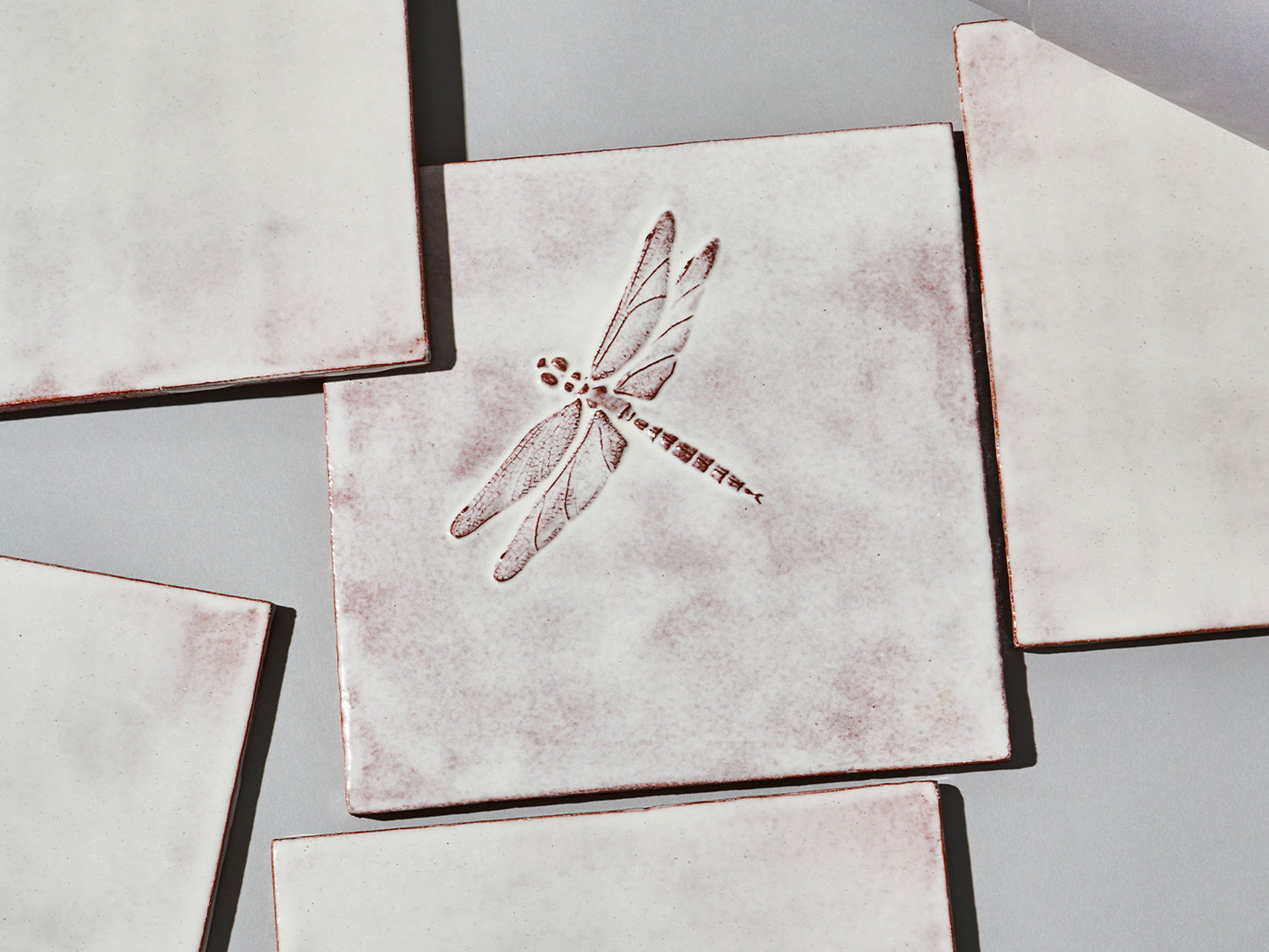 Quadratische, weiße Majolika-Fliese mit leichtem Verlauf und erhabenem Relief einer filigranen Libelle im Zentrum.

Square white-coloured majolica tile with subtle shading and a raised relief of a delicate damselfly in the centre.