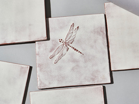 Quadratische, weiße Majolika-Fliese mit leichtem Verlauf und erhabenem Relief einer filigranen Libelle im Zentrum.

Square white-coloured majolica tile with subtle shading and a raised relief of a delicate damselfly in the centre.